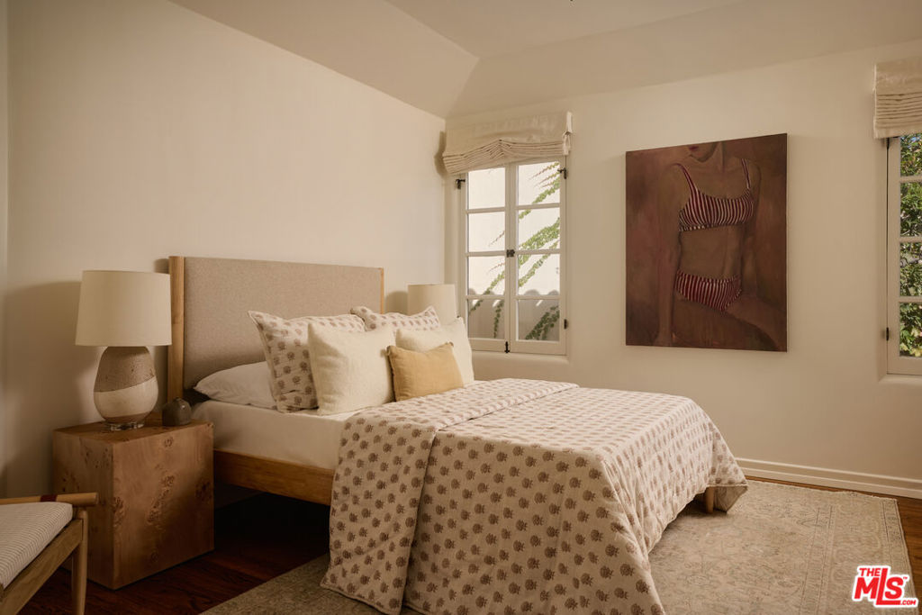 Bedroom with a patterned bedspread, wooden nightstand, and artwork between two windows.