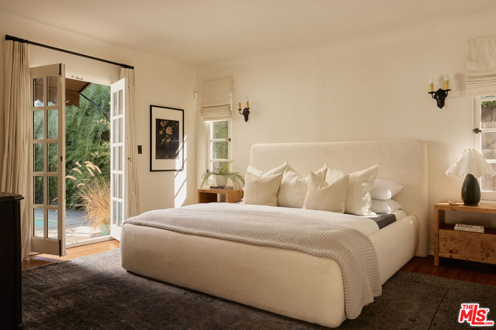 Cream upholstered bed in a bright bedroom with open French doors to greenery and a pool.