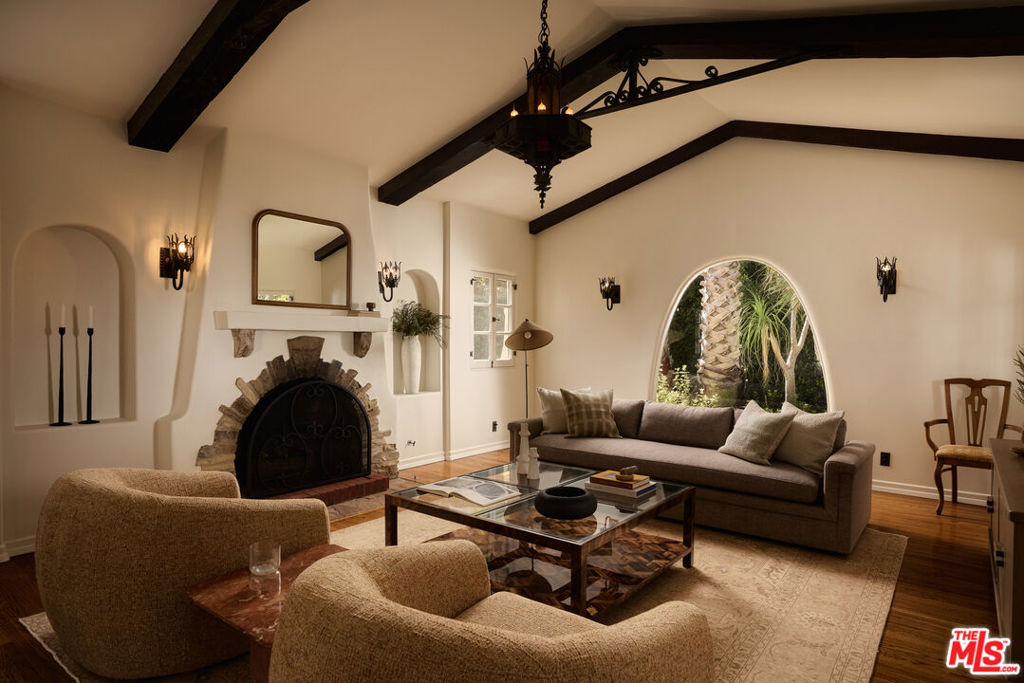 Living room with exposed beams, a stone fireplace, and an arched window overlooking greenery.