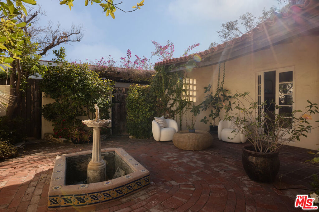 Brick courtyard with a tiled fountain basin, white chairs, and flowering vines beside the house.