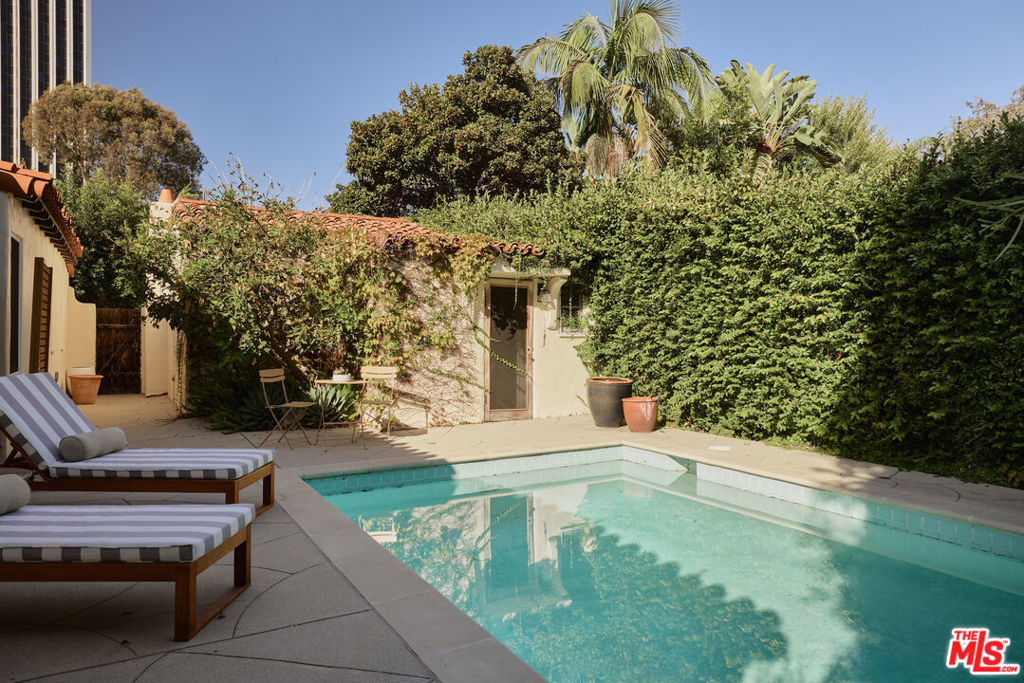 Backyard swimming pool beside a paved patio with striped lounge chairs and tall hedges.
