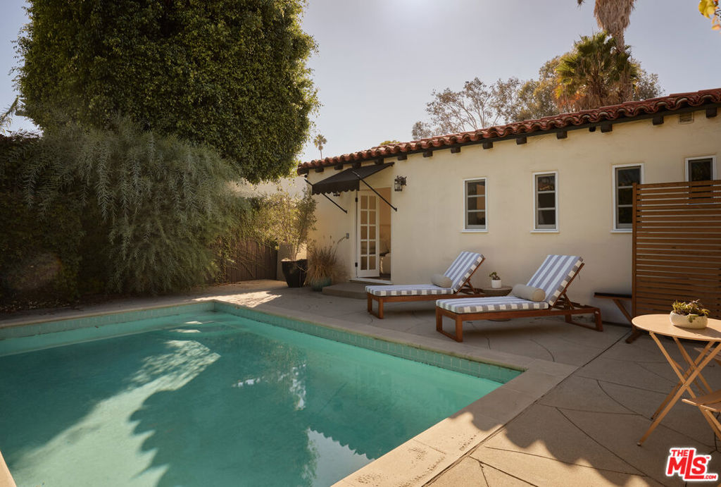 Backyard pool beside a cream stucco home, with striped loungers and mature trees.