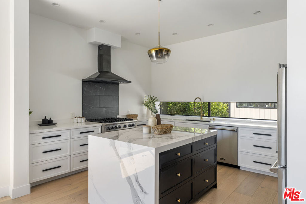 Modern kitchen with a dark island, marble counters, black range hood, and long window overlooking greenery.