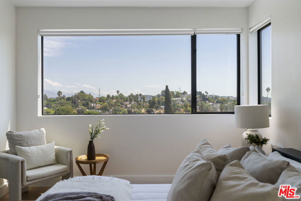 Furnished bedroom with corner windows overlooking a leafy neighborhood and distant hills.