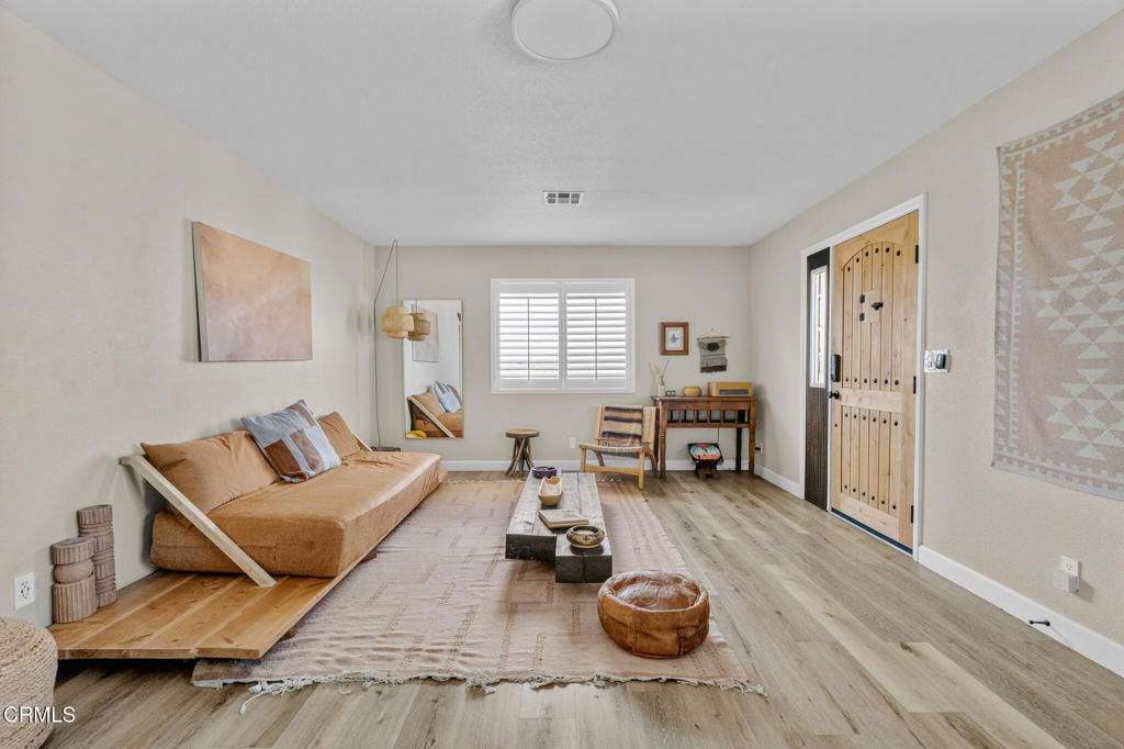 Living room with a raised daybed platform, pale rug, shuttered windows, and a wooden front door.
