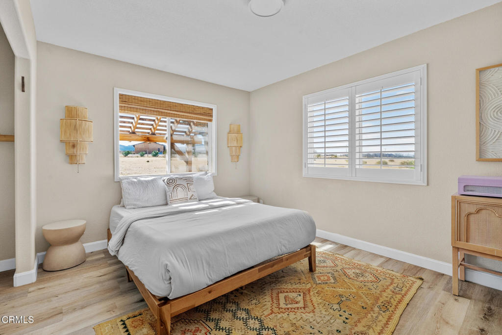 Staged bedroom with a low wooden bed, patterned rug, and shuttered windows overlooking an arid landscape.