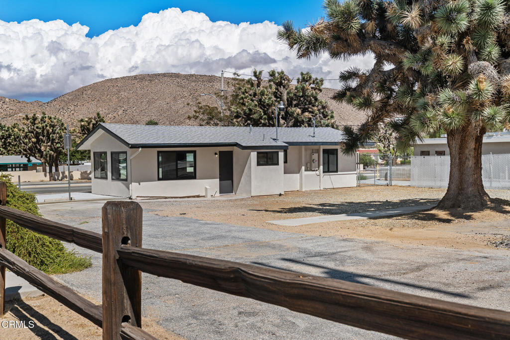 Single-story building with a gray roof, framed by a wooden rail and Joshua tree, with desert hills behind it.