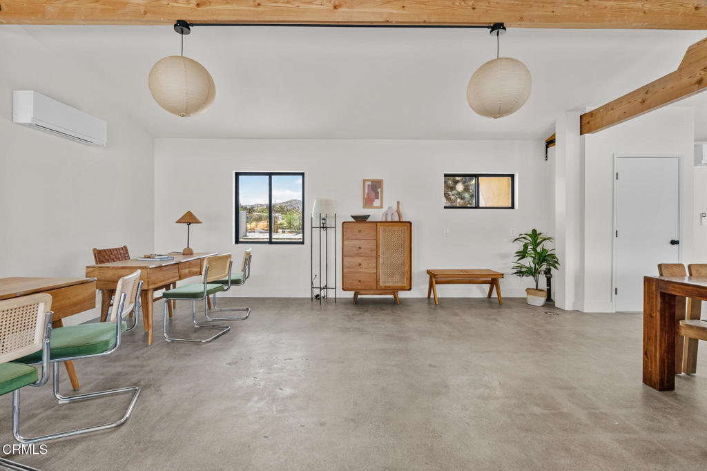 Dining area with a wooden table, green chairs, concrete floor, and windows overlooking desert hills.