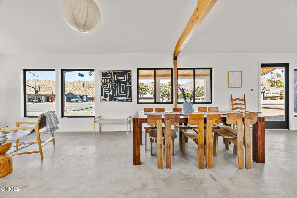 Dining area with a large wooden table, exposed ceiling beam, and windows overlooking desert hills.