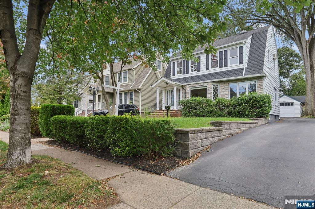 Two-story white house with stone facade, dark shutters, landscaped hedges, and a driveway.