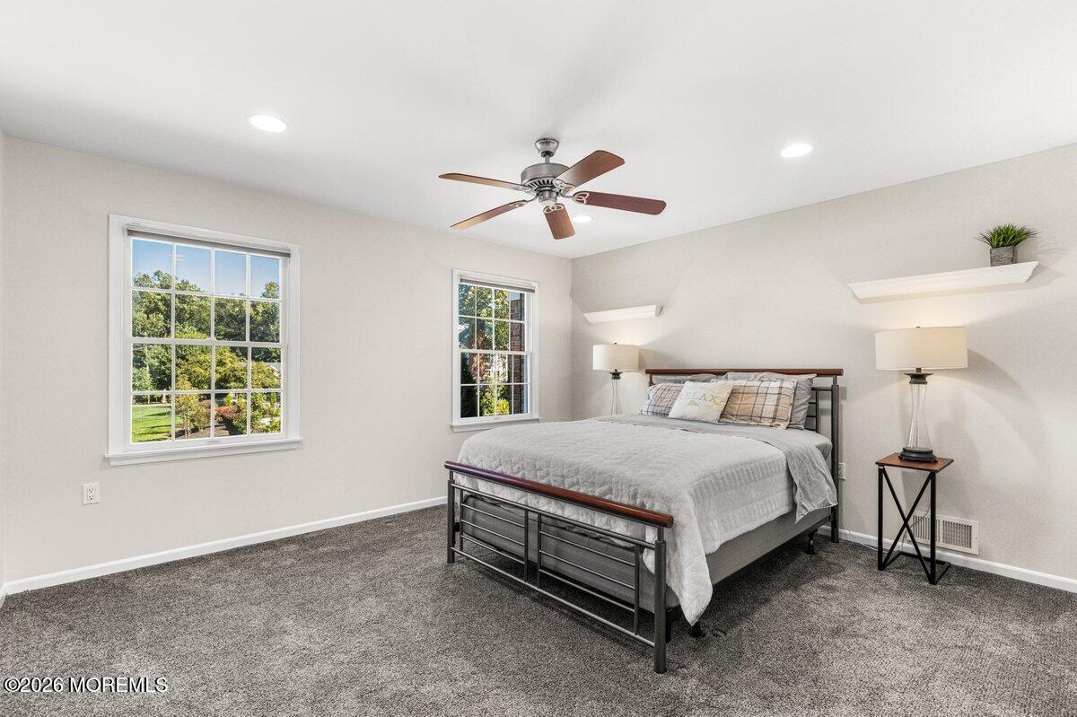 Bedroom with a metal-frame bed, gray carpet, and two windows overlooking leafy greenery.