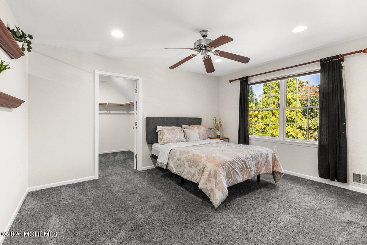 Bedroom with a patterned bedspread, gray carpet, ceiling fan, and large curtained window overlooking trees.
