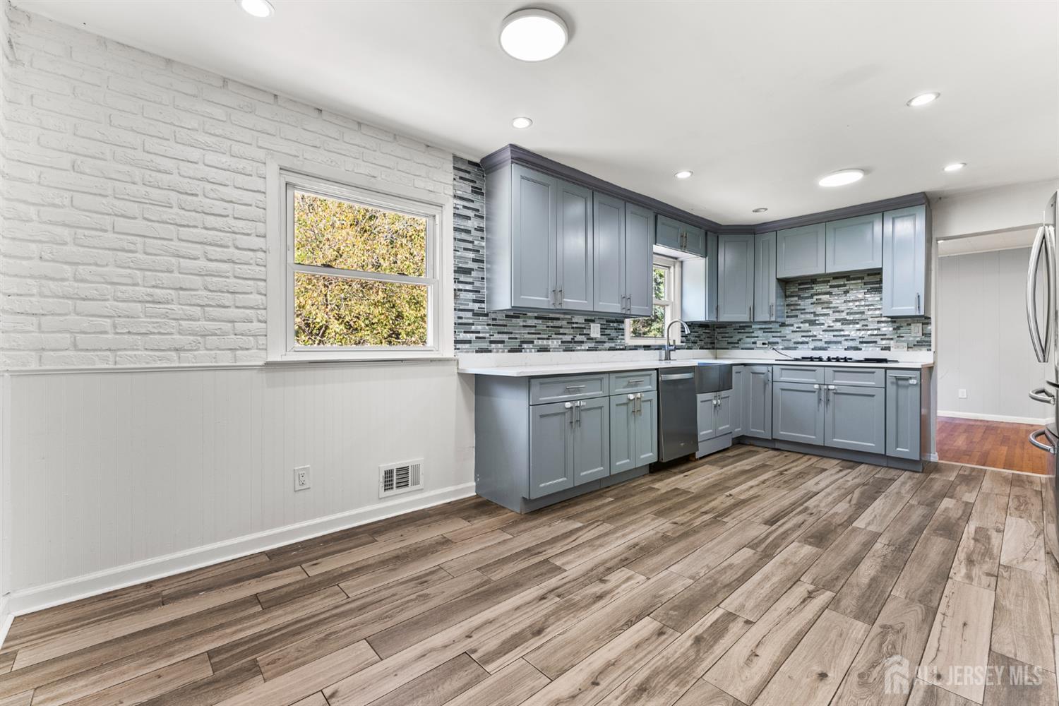 Renovated kitchen with blue-gray cabinets, mosaic backsplash, white brick wall, and wood-look floors.
