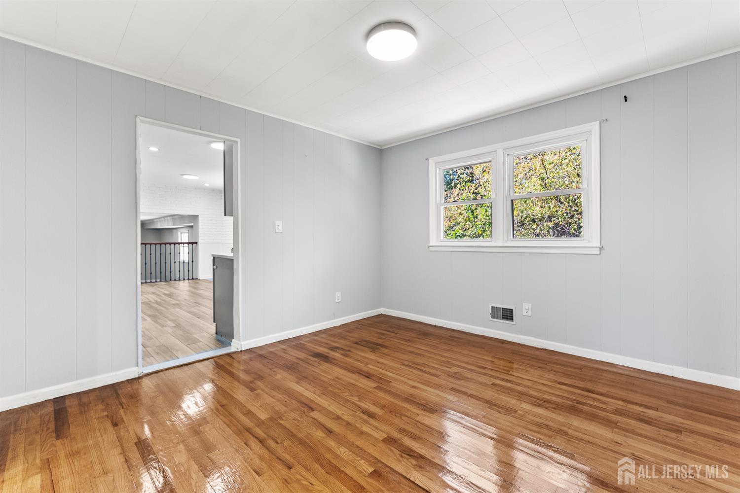 Vacant room with polished hardwood floors, gray paneled walls, and a double window overlooking trees.