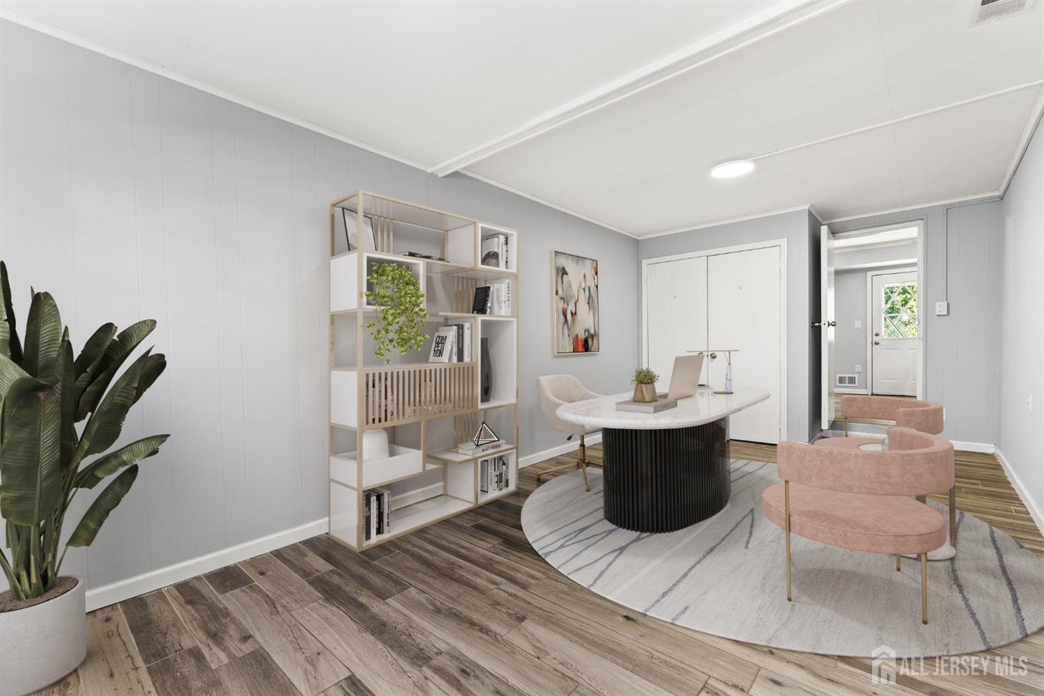 Staged home office with an oval desk, pink chairs, open shelving, and wood-look flooring.