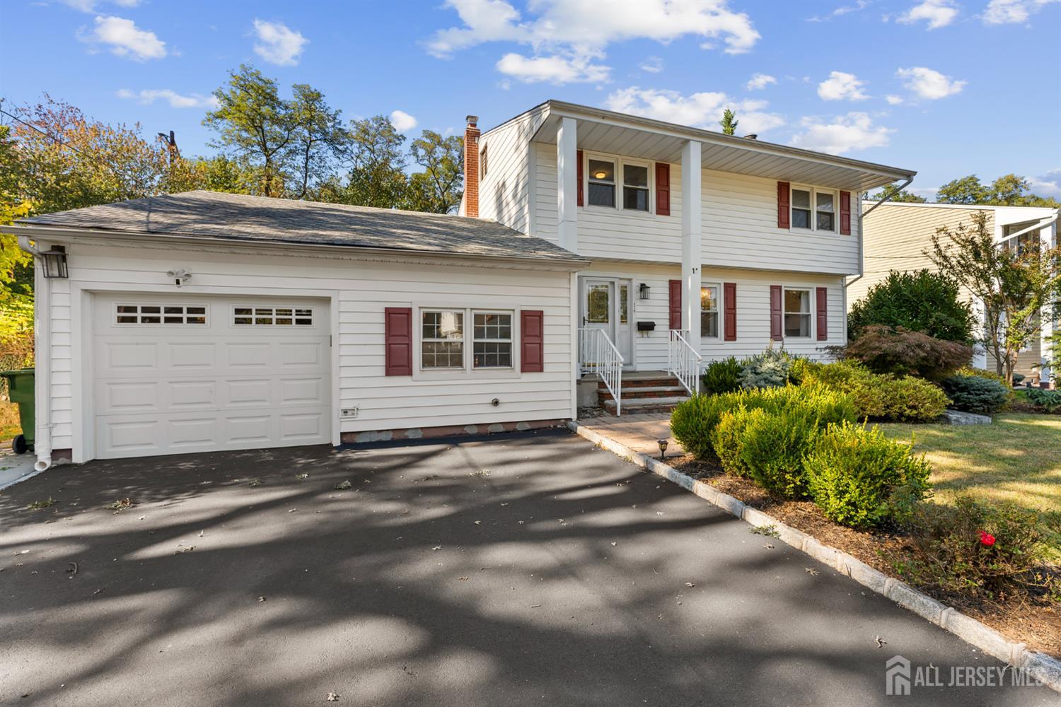 White split-level house with red shutters, an attached garage, landscaped shrubs, and a wide paved driveway.