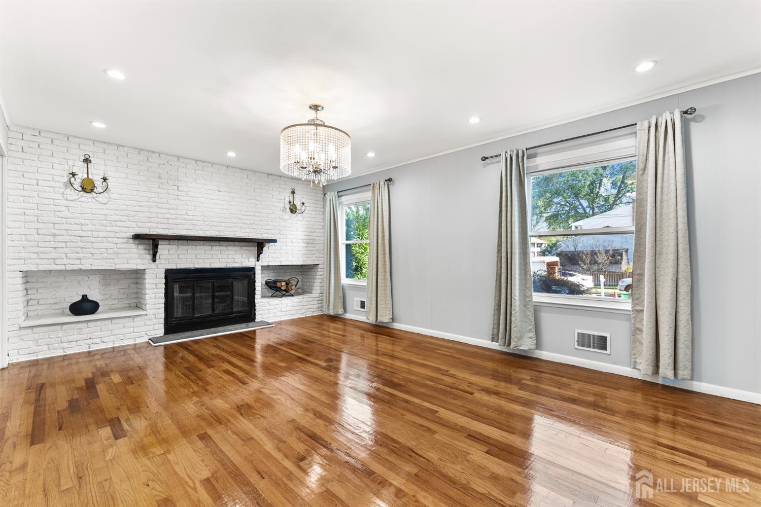Living room with a white brick fireplace, polished hardwood floors, and curtained windows.