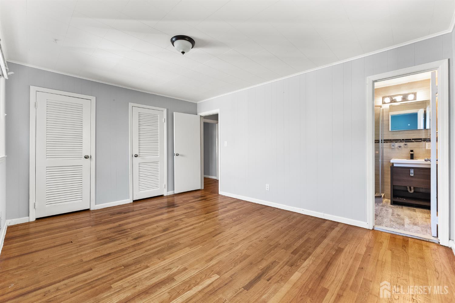 Empty bedroom with hardwood floors and louvered closet doors; adjoining bathroom visible through the doorway.