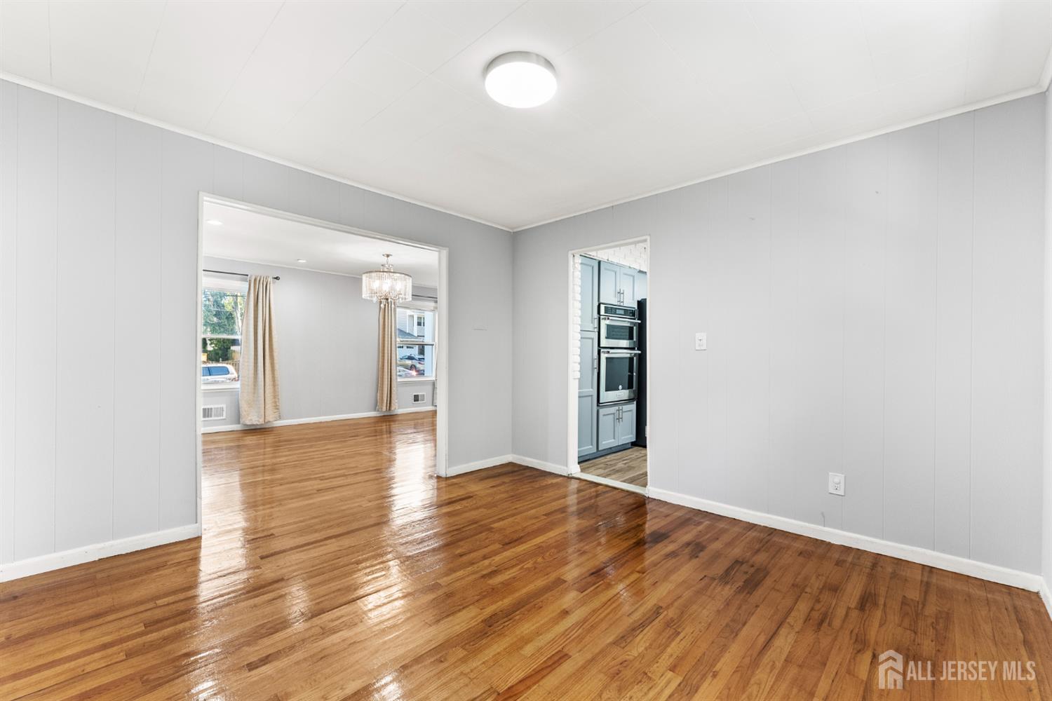 Empty room with glossy hardwood floors, opening to a bright room and blue-gray kitchen.