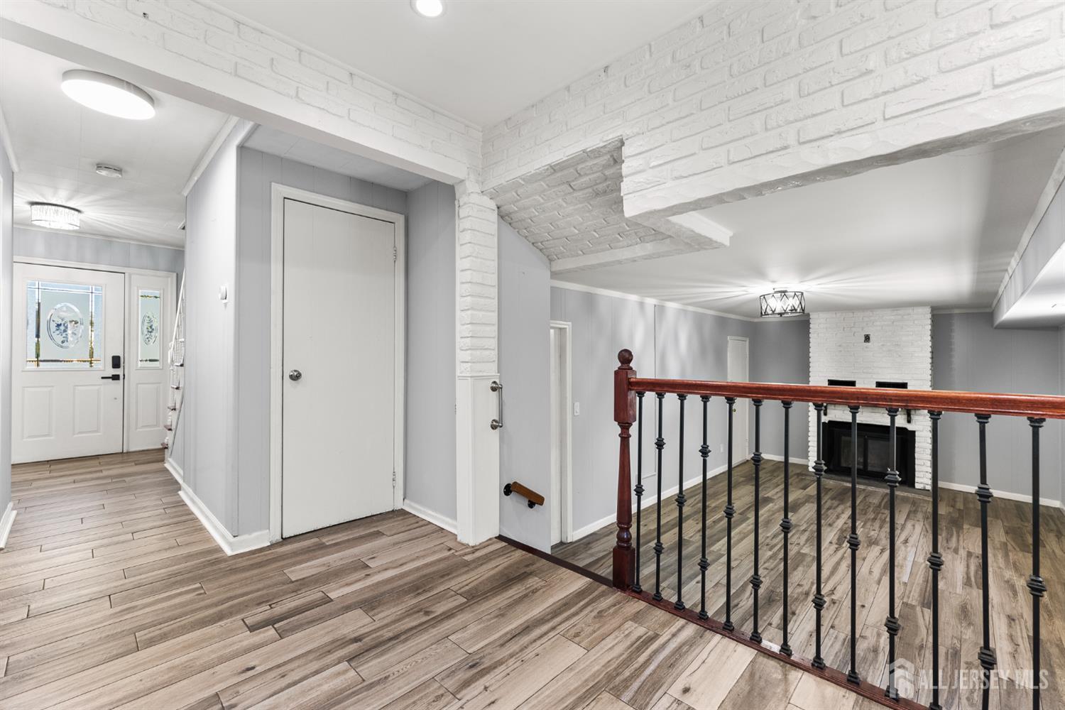 Entry hall opening to a living area with wood-look tile, a dark railing, and a white brick fireplace.