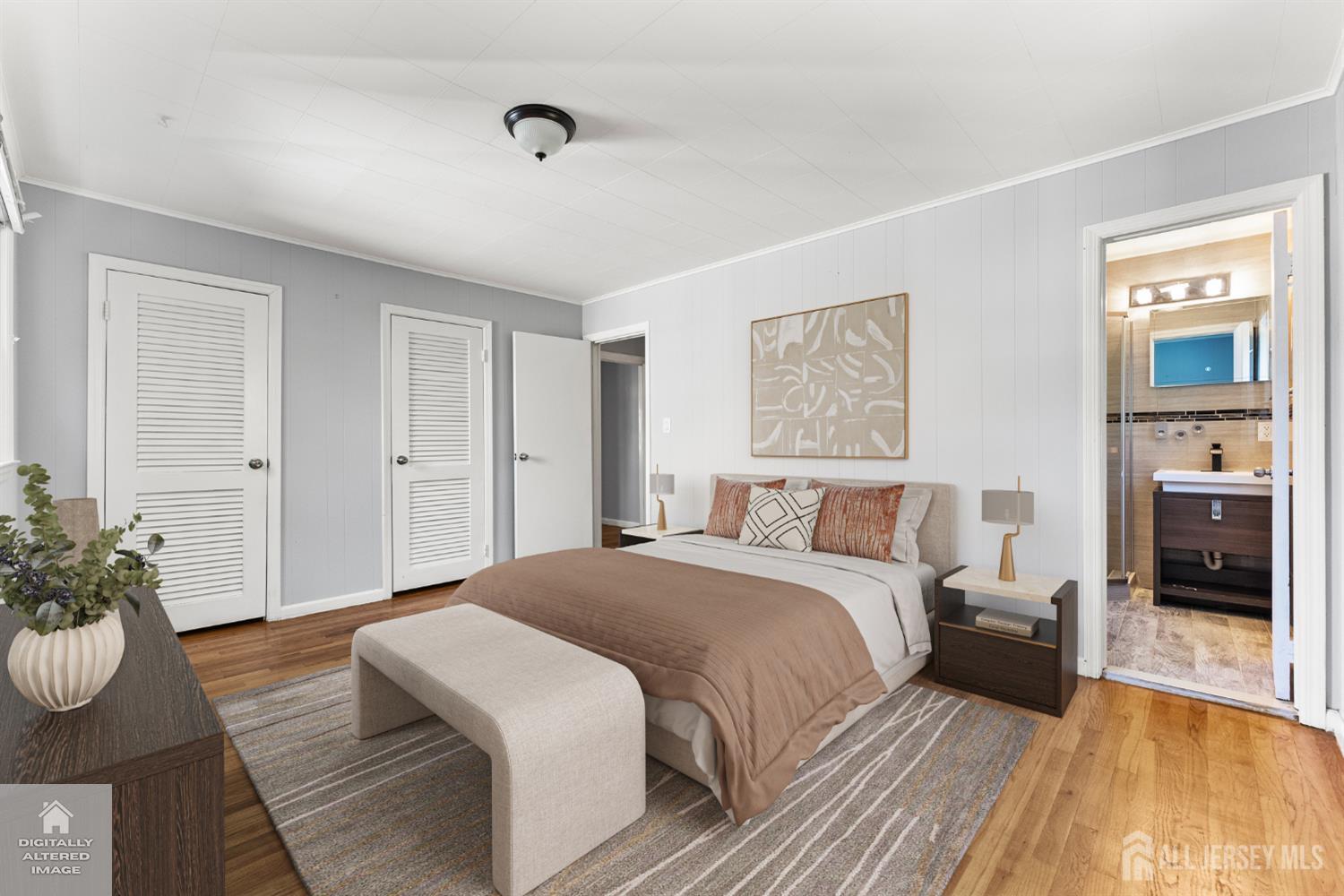 Staged bedroom with taupe bedding, an upholstered bench, and an adjoining bathroom.