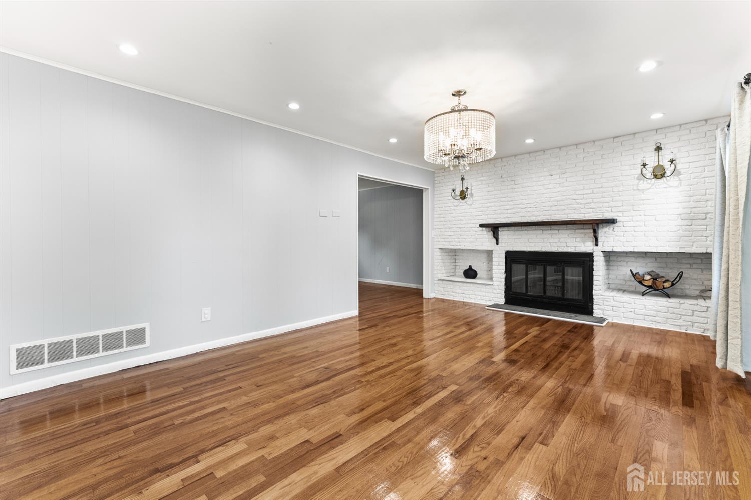 Empty living room with hardwood floors, a white brick fireplace, and a crystal chandelier.
