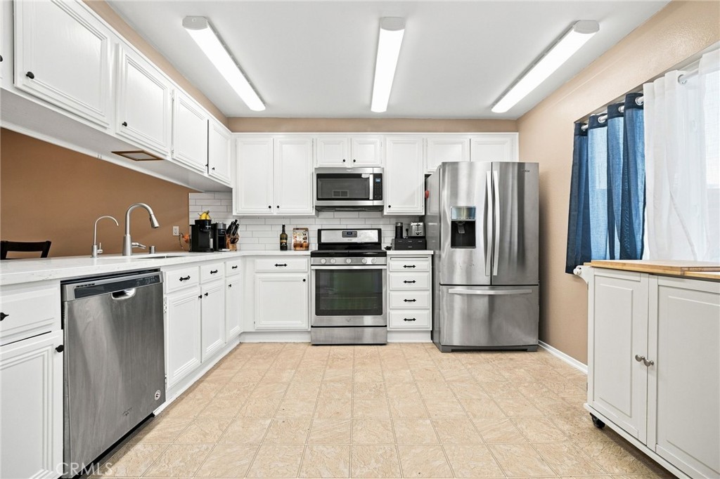 Kitchen with white cabinets, stainless steel appliances, beige tile floors, and blue curtains.