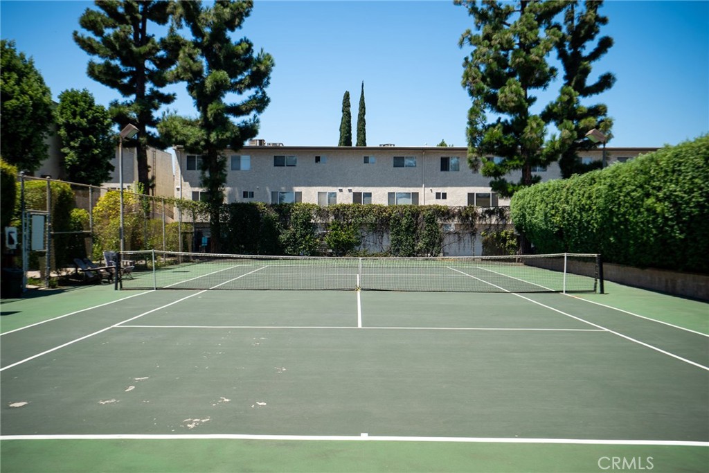 Outdoor tennis court with a net, tall trees, and a residential building behind it.