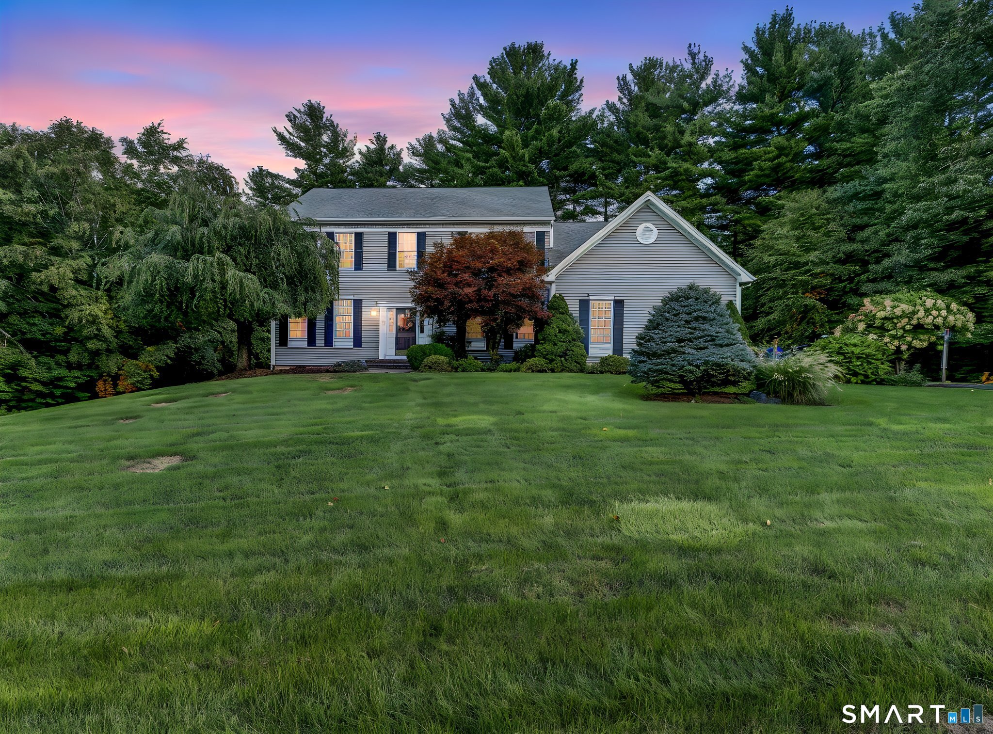 Gray two-story home with blue shutters, mature trees, and a broad lawn at dusk.