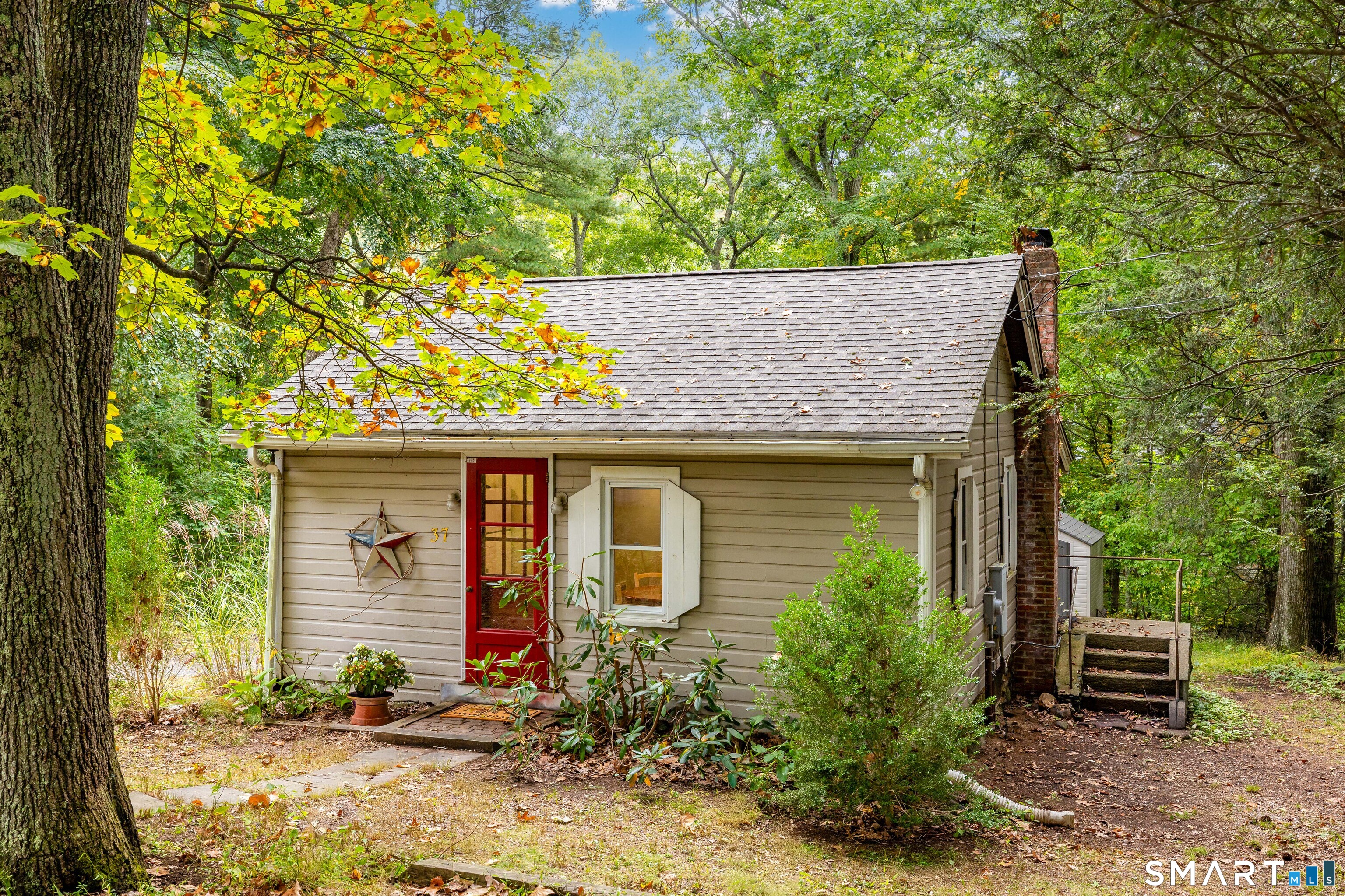Cottage among mature trees, with a red front door, gray shingle roof, and brick chimney.