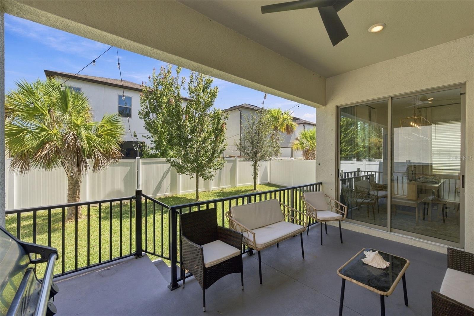 Covered patio with cushioned seating, black railing, and sliding glass doors overlooking a fenced lawn.