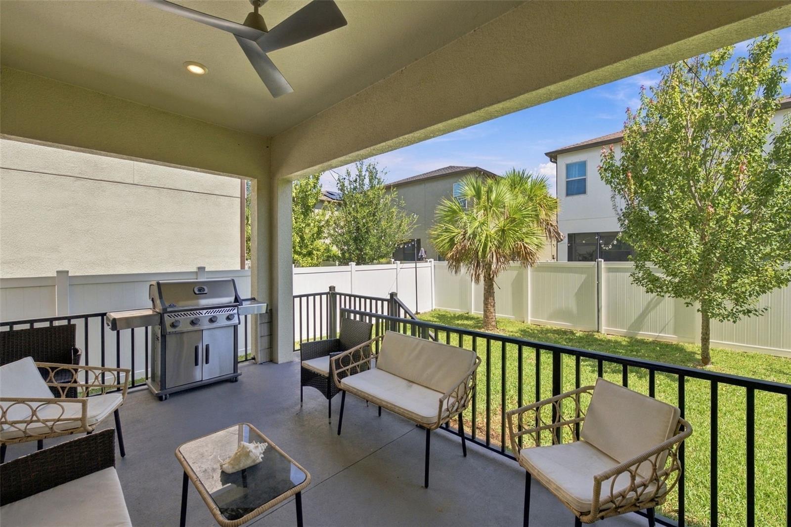 Covered patio with wicker seating and a stainless steel grill overlooking a fenced, tree-lined yard.