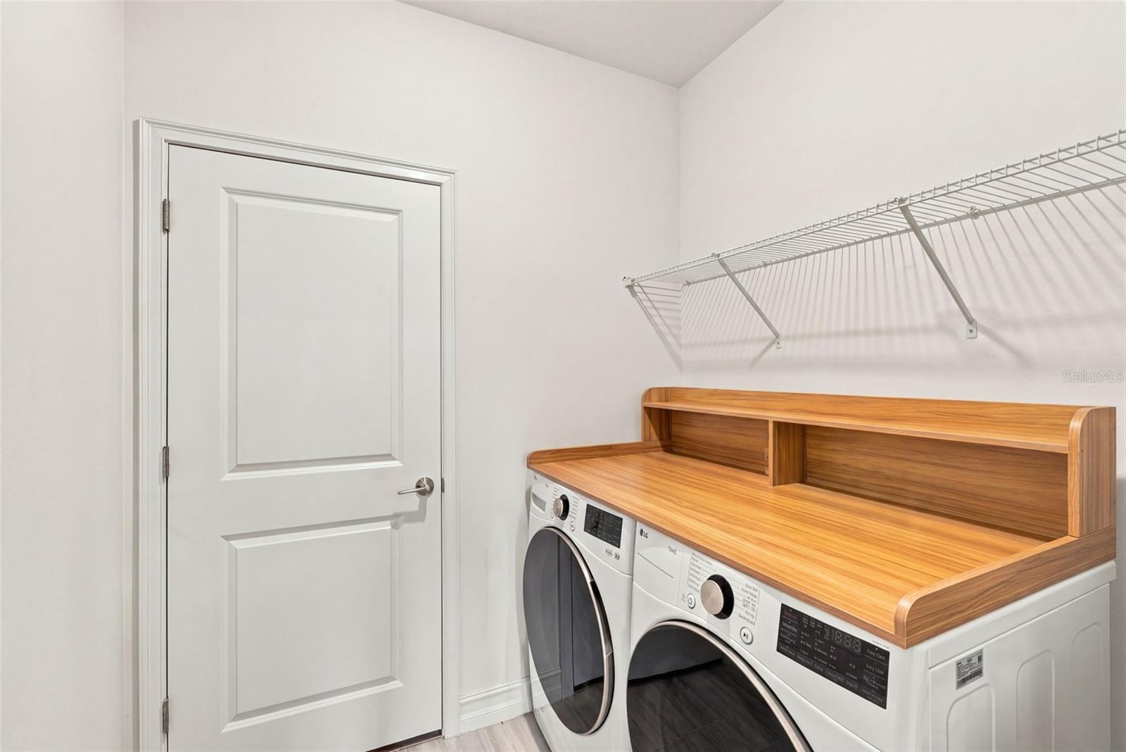 Laundry room with front-loading washer and dryer beneath a wood countertop and wall-mounted wire shelf.