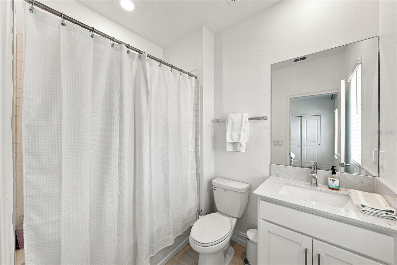 White bathroom with a curtained shower, toilet, and vanity topped with a large mirror.
