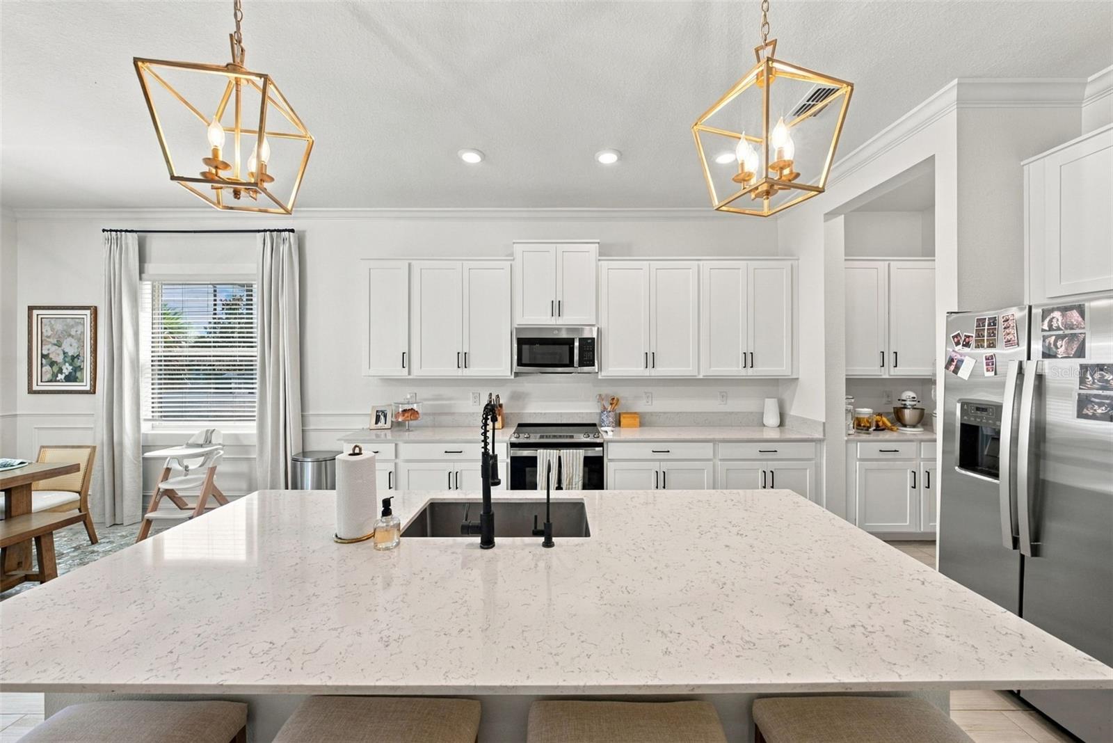 Bright white kitchen with a large quartz island, dark sink, gold pendant lights, and stainless steel appliances.