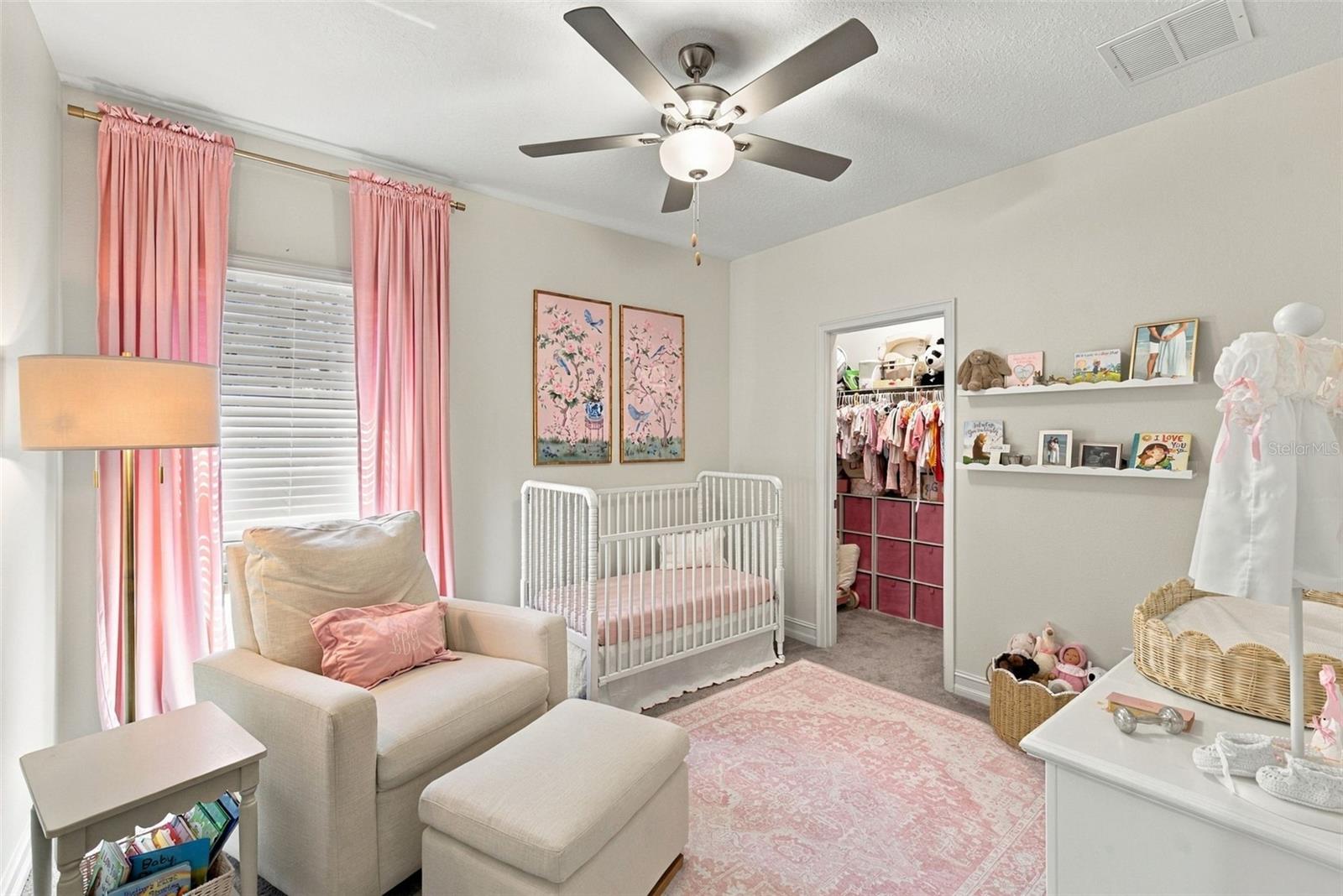 Nursery with a white crib, beige armchair, and pink curtains and rug beside an open closet.
