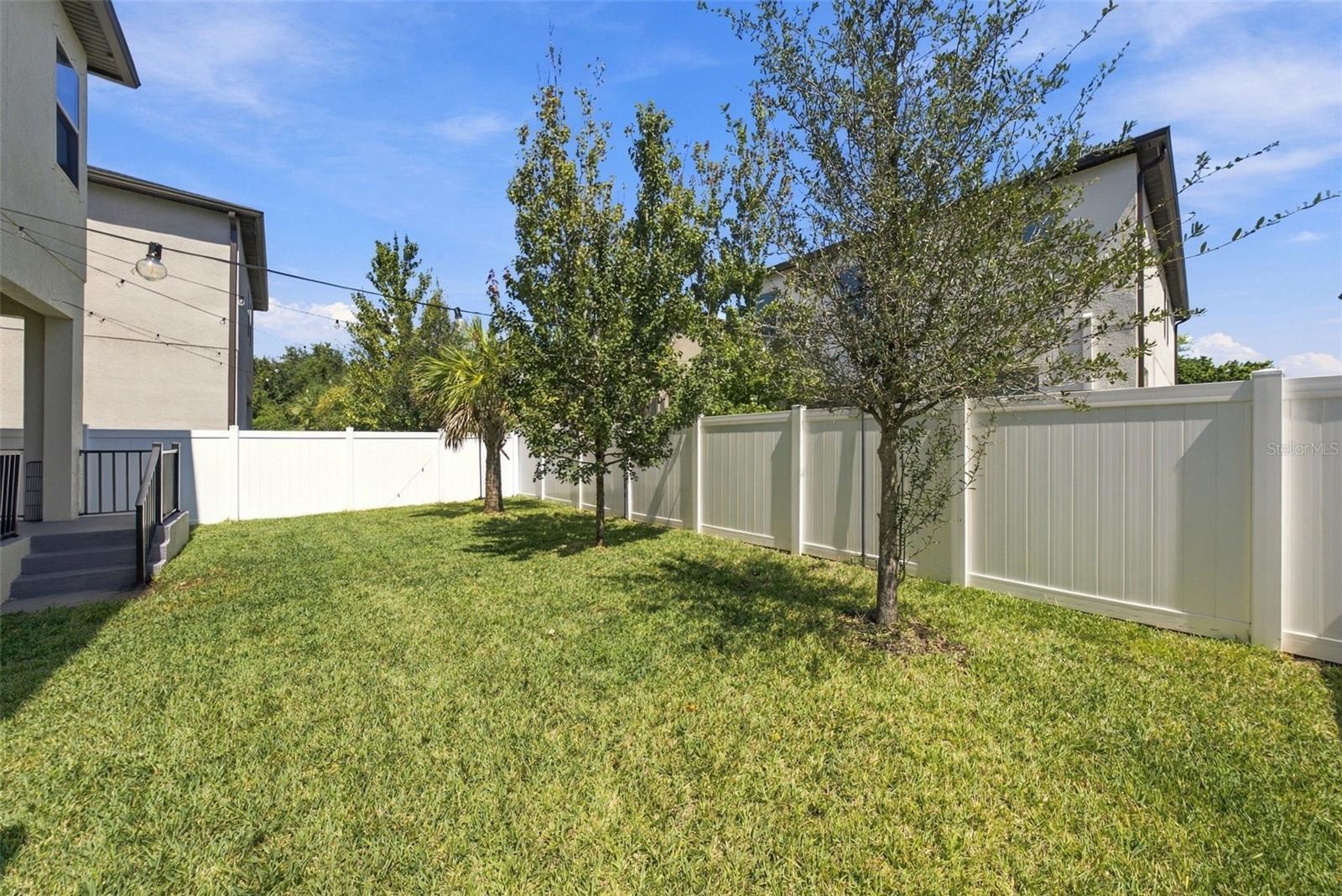 Grassy backyard with a white privacy fence, several trees, and steps to a covered patio.