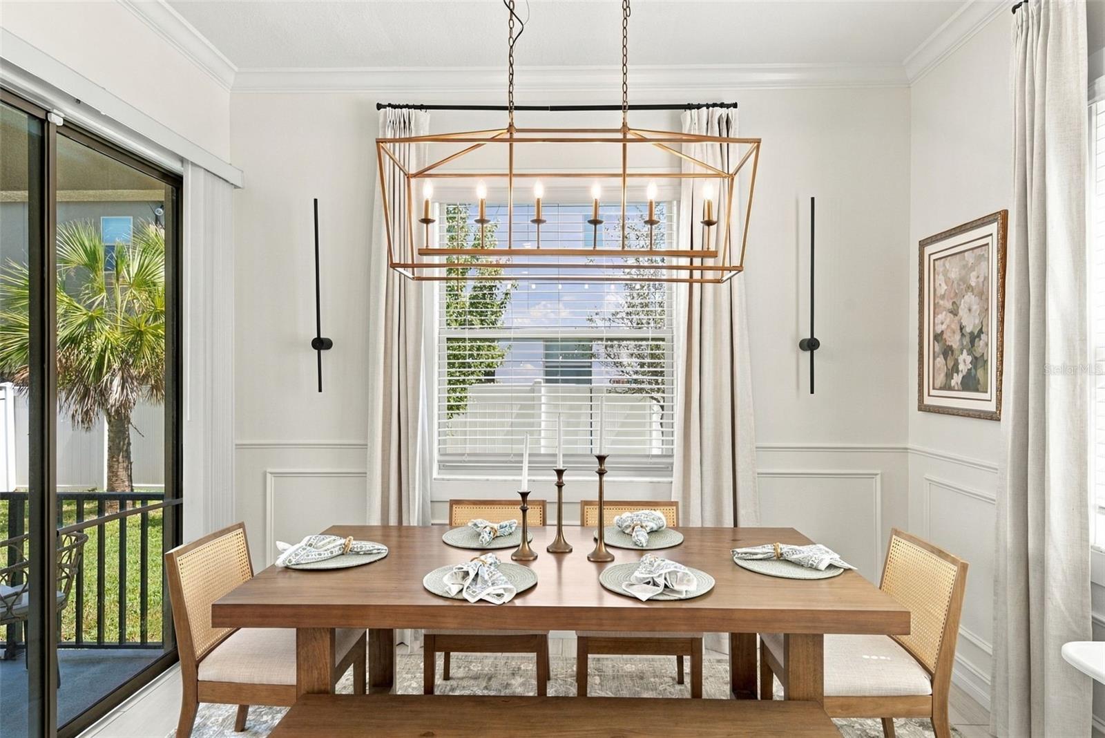 Dining room with a set wooden table beneath a geometric brass chandelier, beside sliding glass doors.