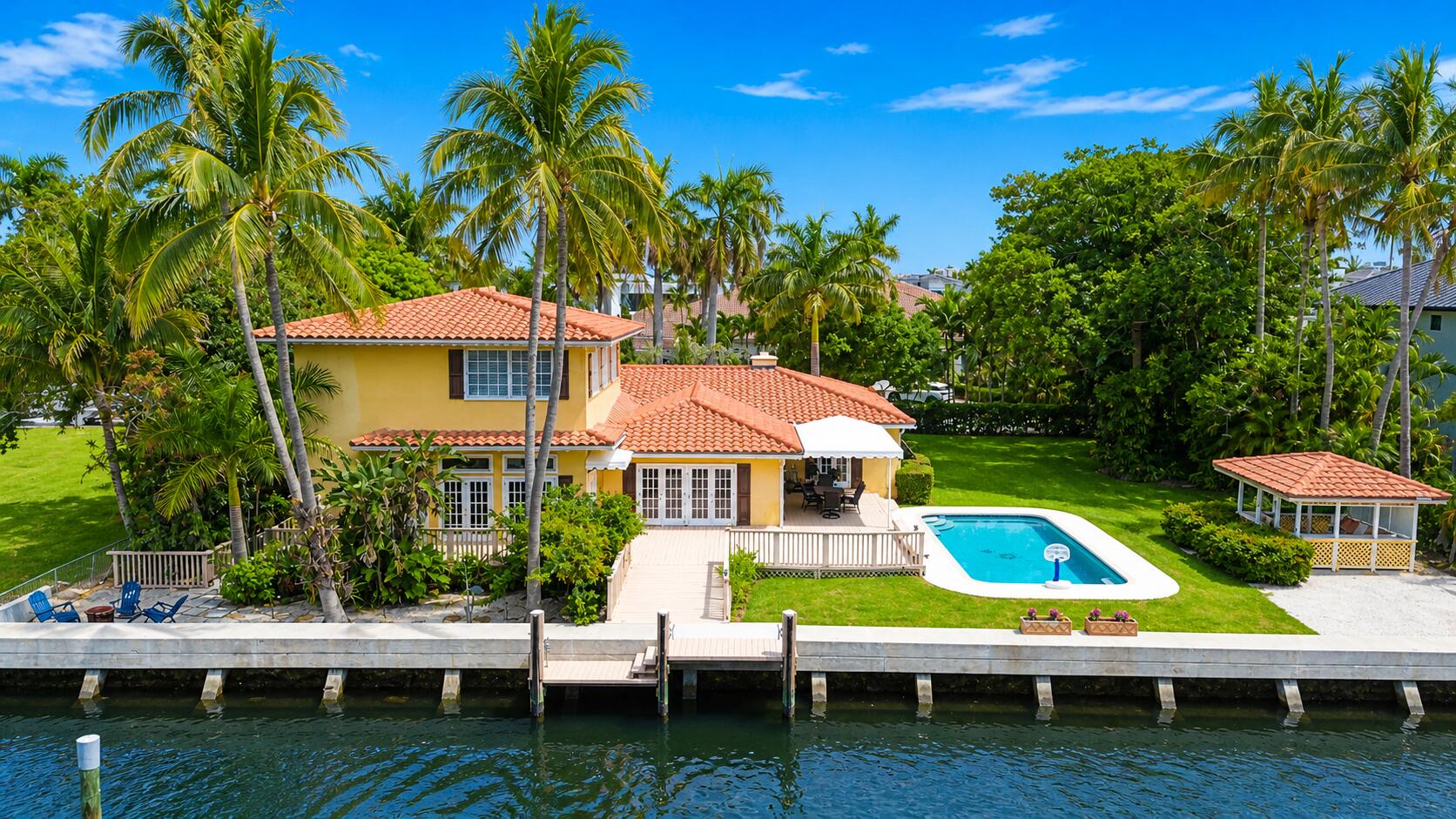 Aerial view of a yellow waterfront home with a pool, canal dock, and palm-lined lawn.