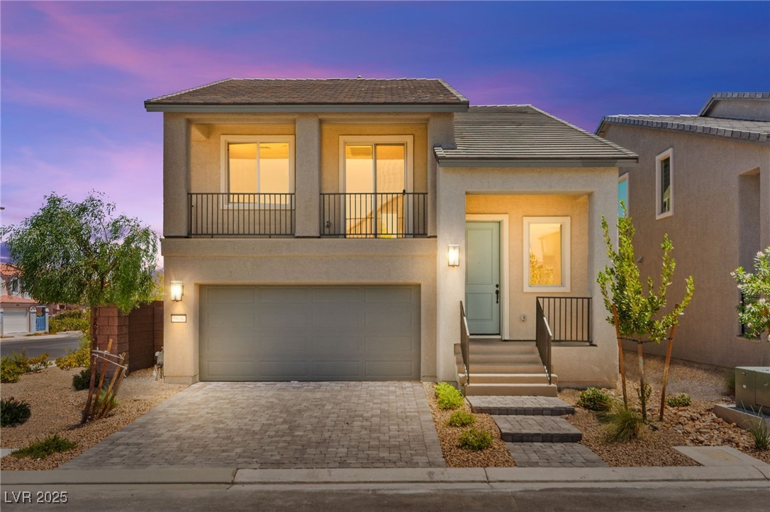Two-story stucco home with upstairs balconies, a double garage, and a paver driveway at dusk.