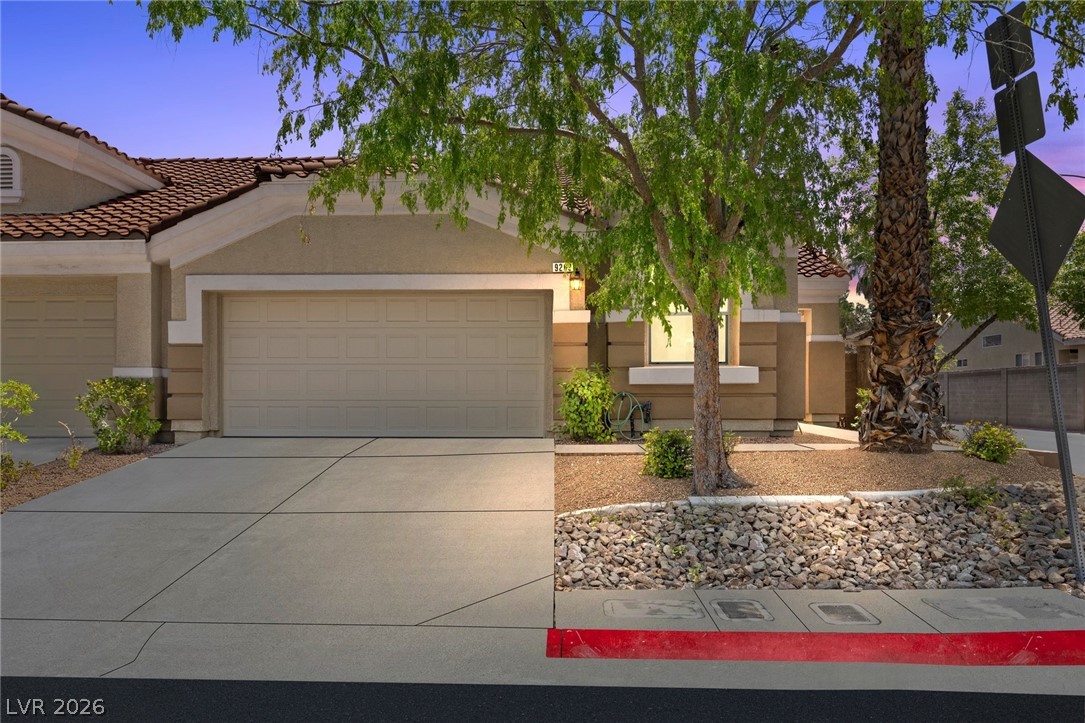 Stucco home with a tiled roof, double garage, and desert landscaping beneath mature trees.