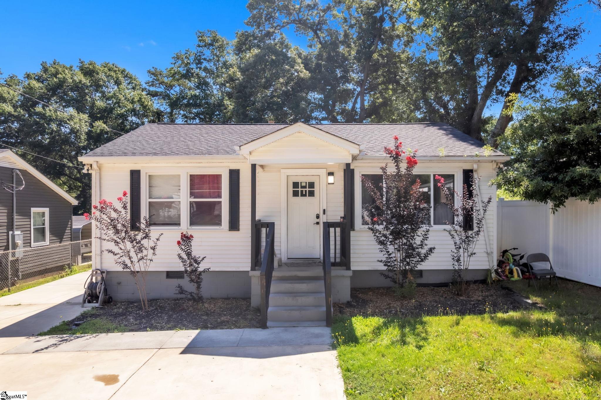 White single-story home with dark shutters, a covered entry, concrete driveway, and mature trees.