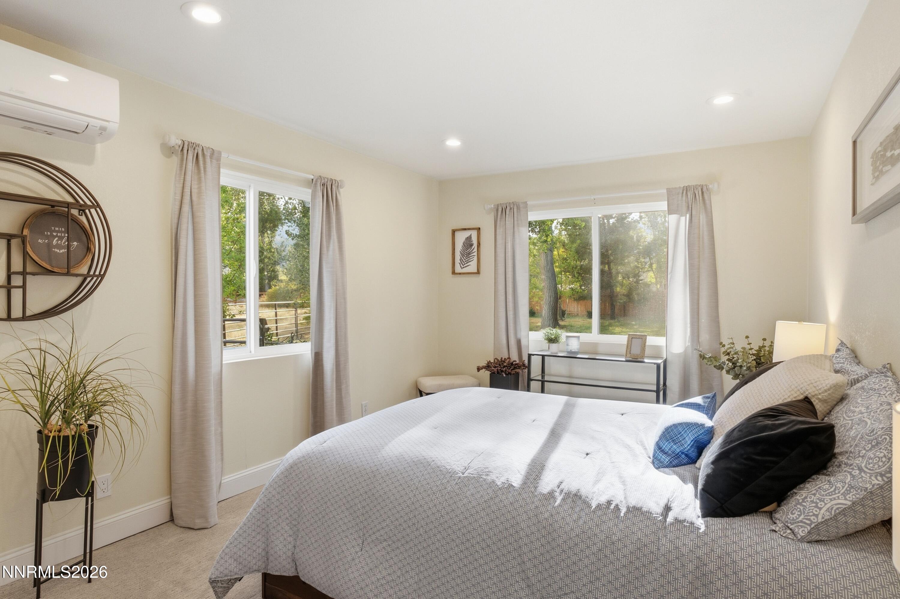 Furnished bedroom with a large gray bed and two windows overlooking leafy trees.