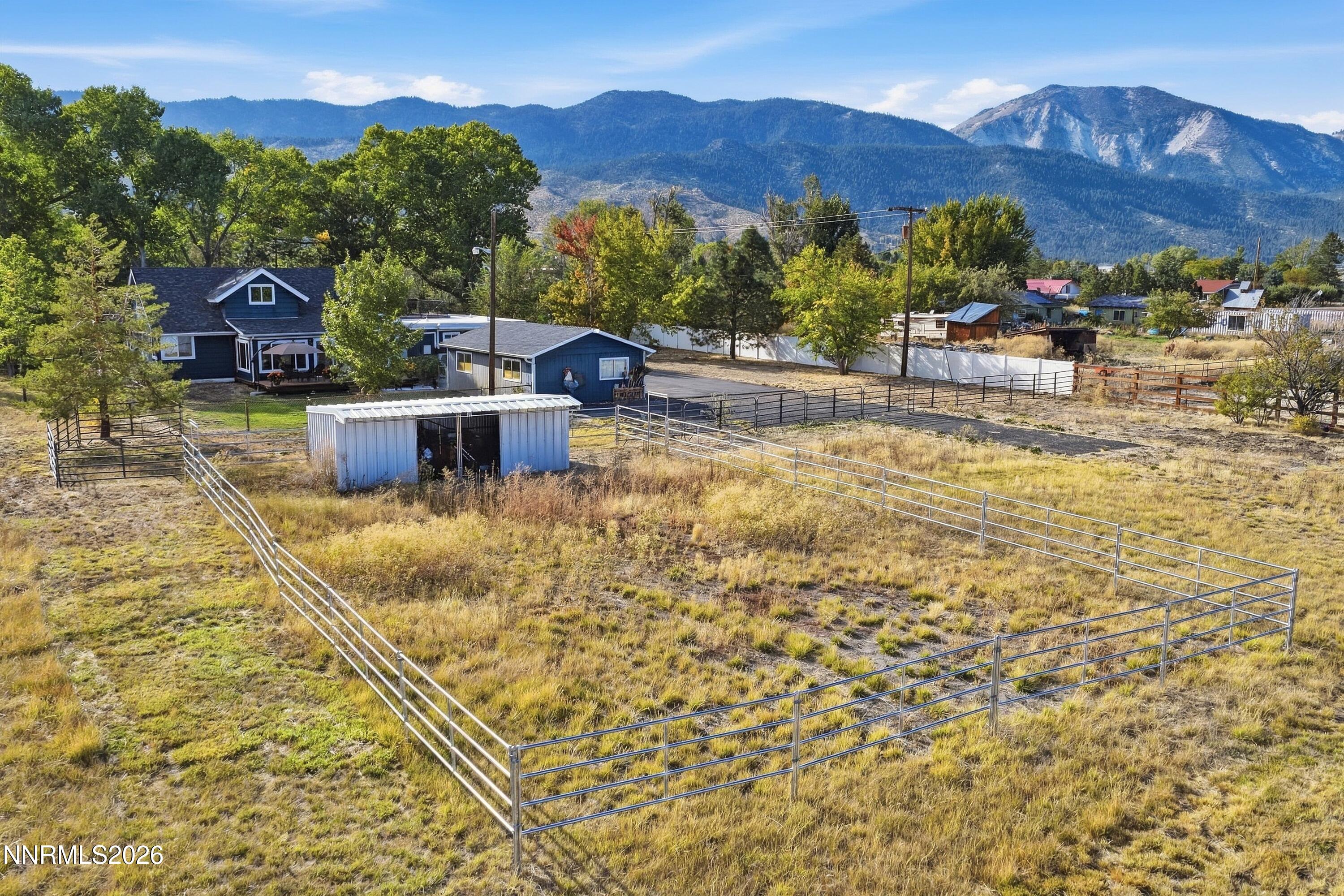 Rural property with fenced paddocks, a blue house, outbuildings, and mountains beyond.
