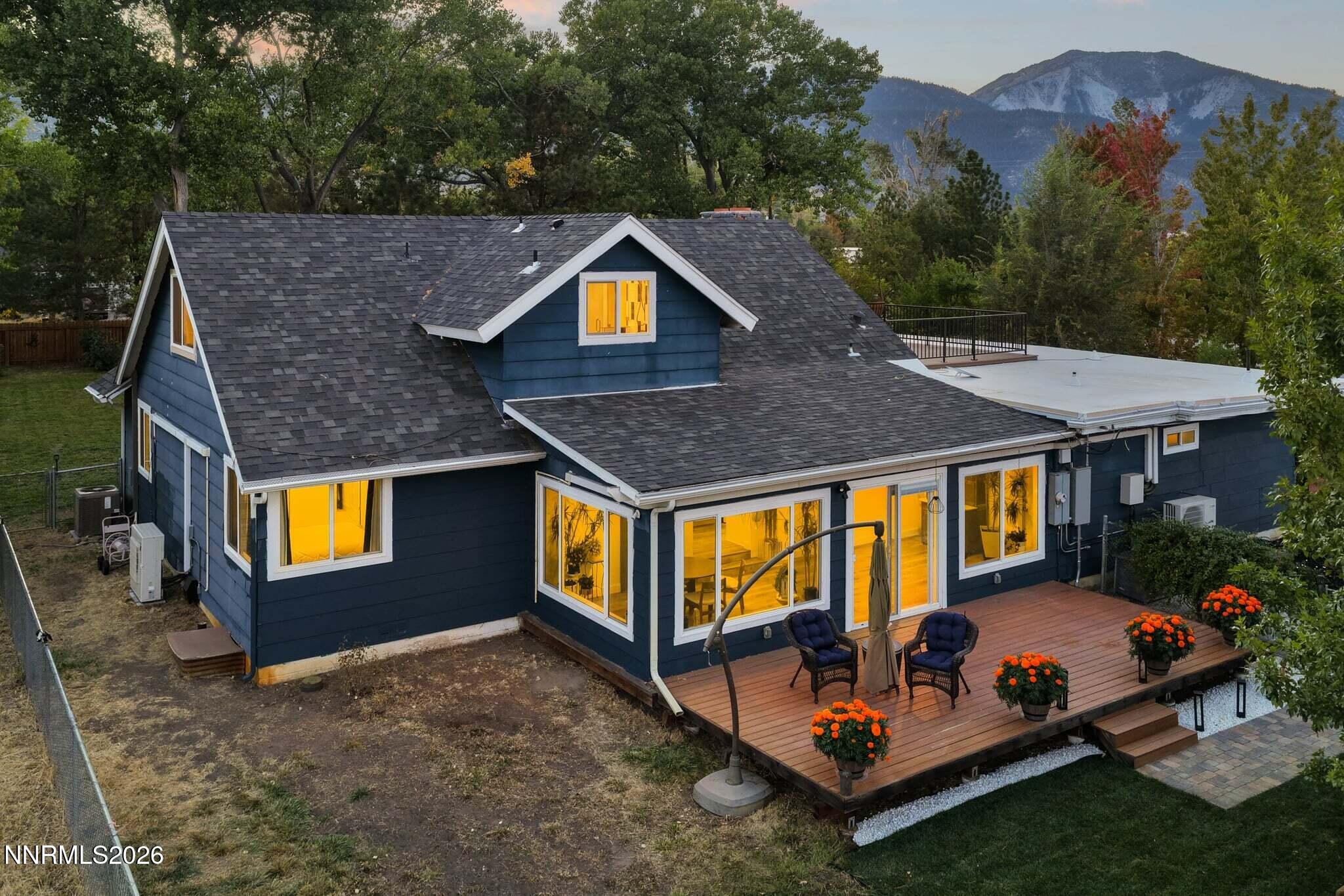 Rear of a dark blue house with a furnished deck, glowing windows, and a mountain backdrop.
