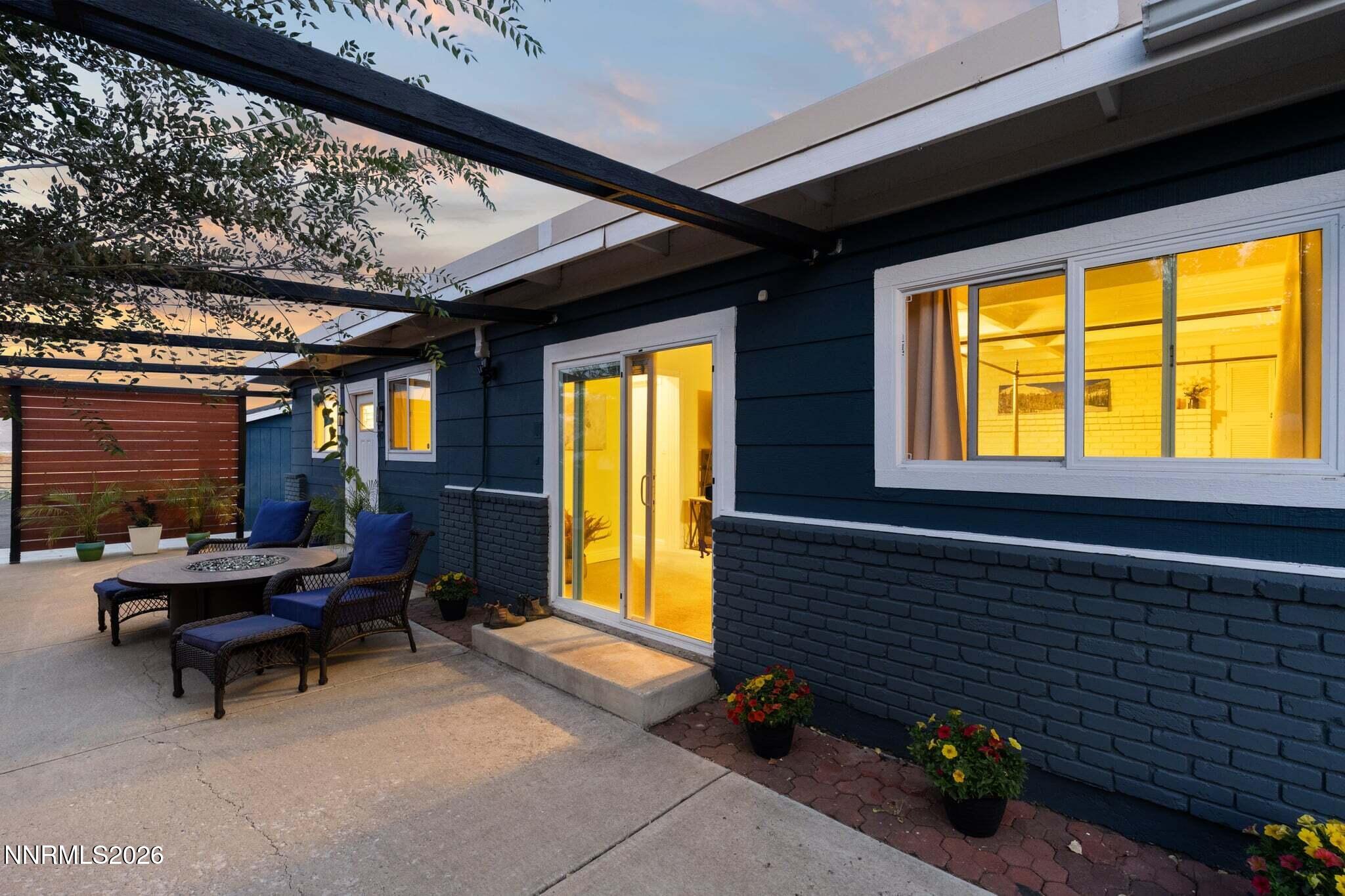 Furnished patio beside a dark blue house, with pergola seating and warmly lit sliding glass doors.
