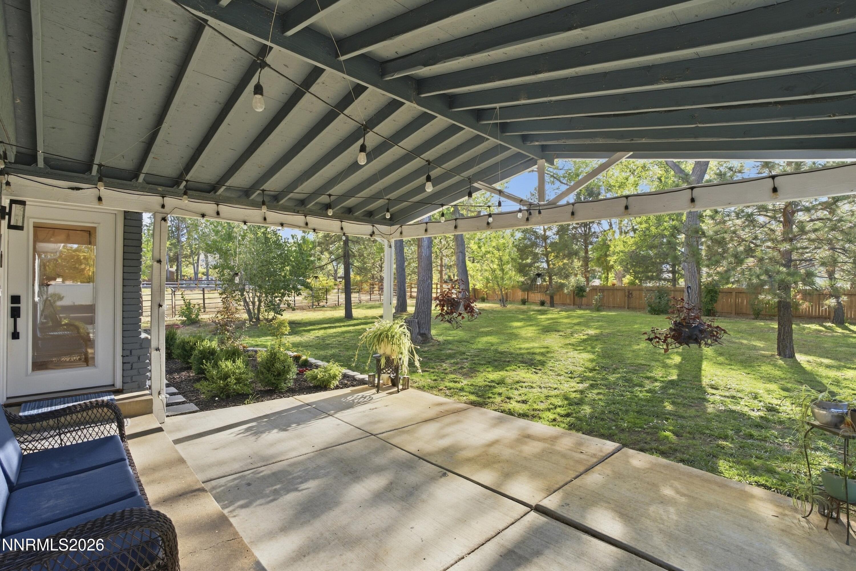 Covered patio with string lights and wicker seating overlooking a fenced, tree-lined lawn.