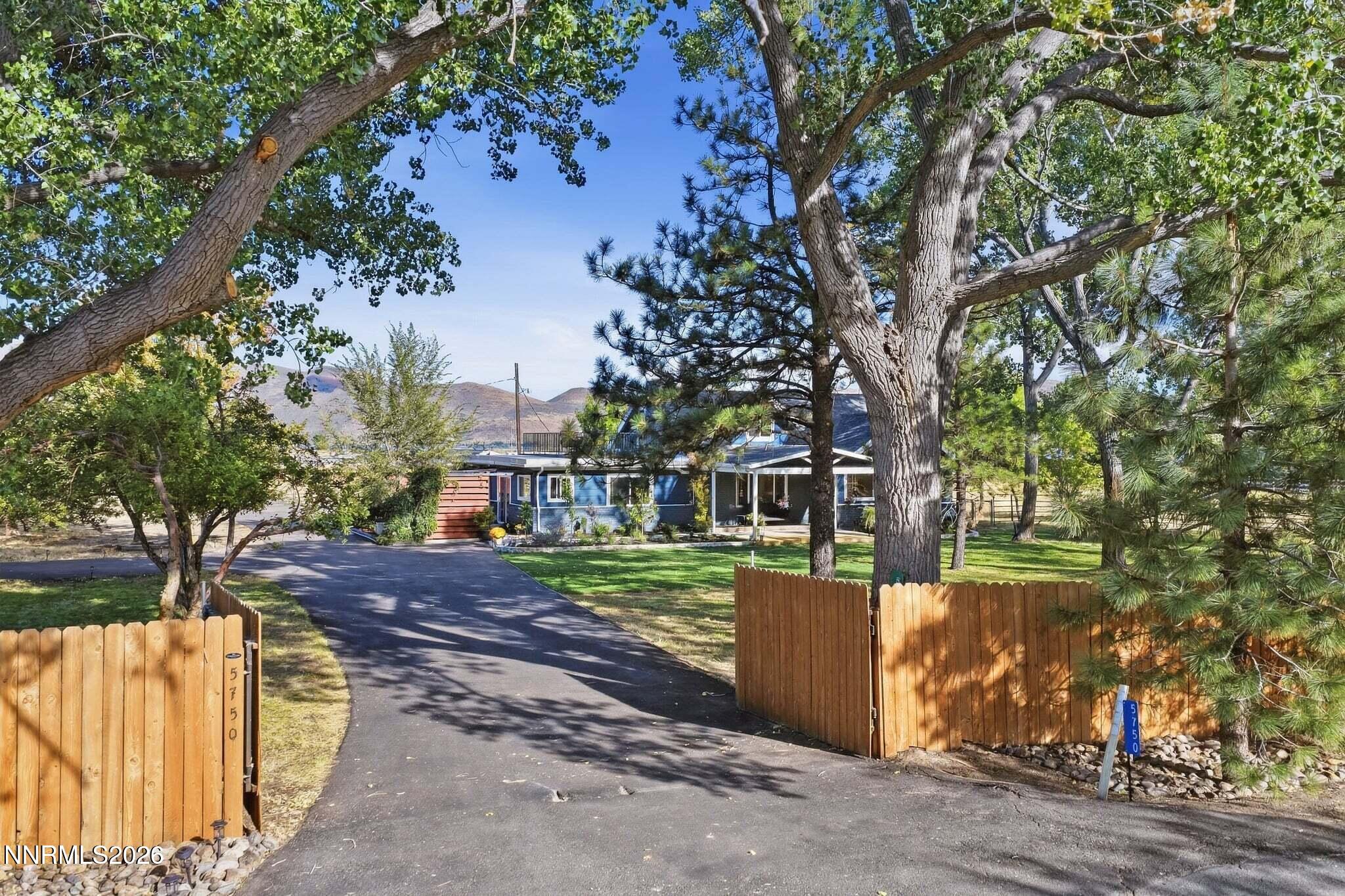 Blue home behind a wooden fence, reached by a paved driveway beneath mature trees.