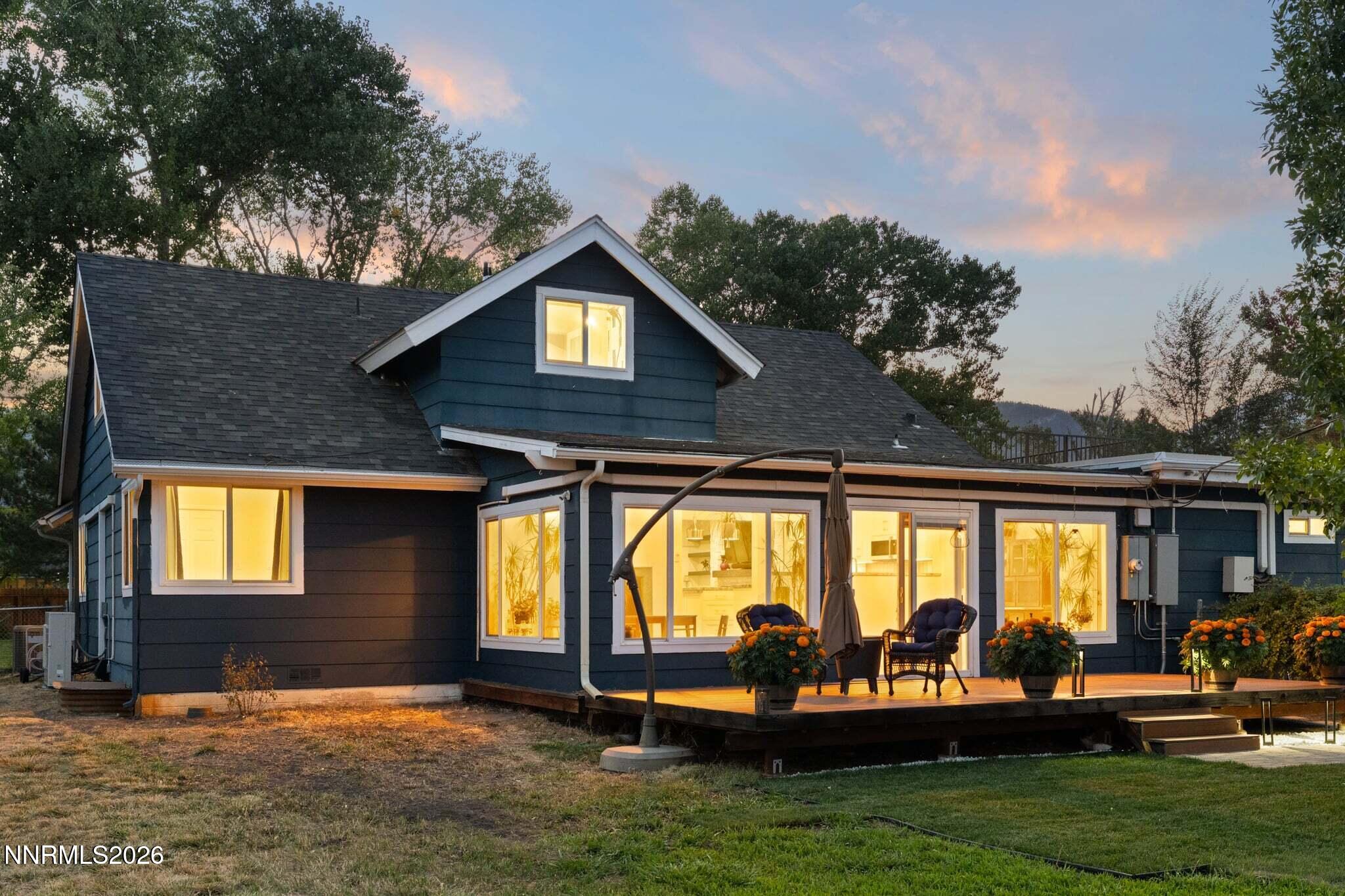 Dark blue house at dusk with a warmly lit rear patio, outdoor chairs, and potted flowers.