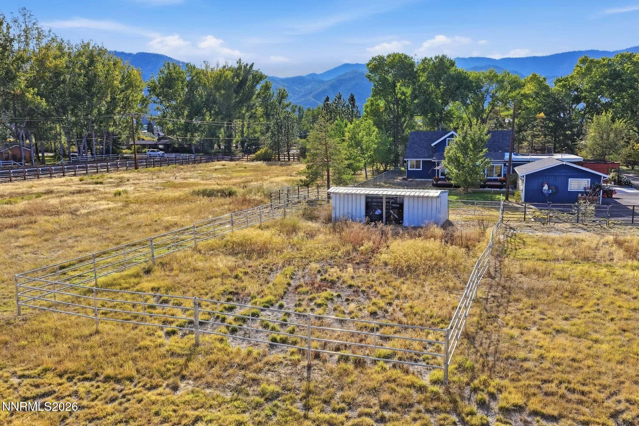 Aerial view of a fenced property with a blue house, outbuildings, and mountain views.